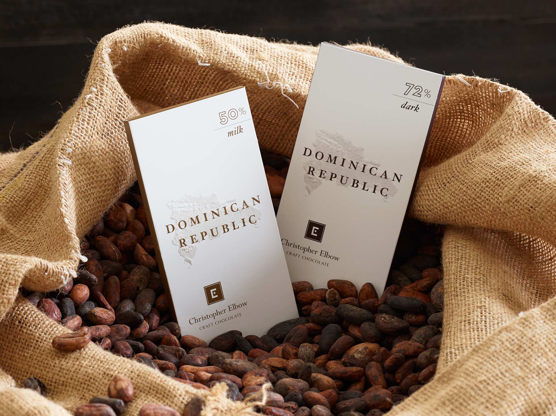 Dark and milk Dominican Republic Christopher Elbow Craft Chocolate bars sitting in an open burlap bag of cocoa beans.