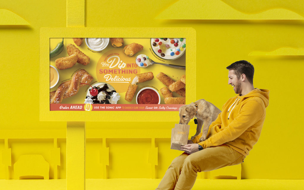 Stylized SONIC drive-in with Man and his puppy in front of POP showing many snack items that says "That Moment You Dip into Something Delicious"