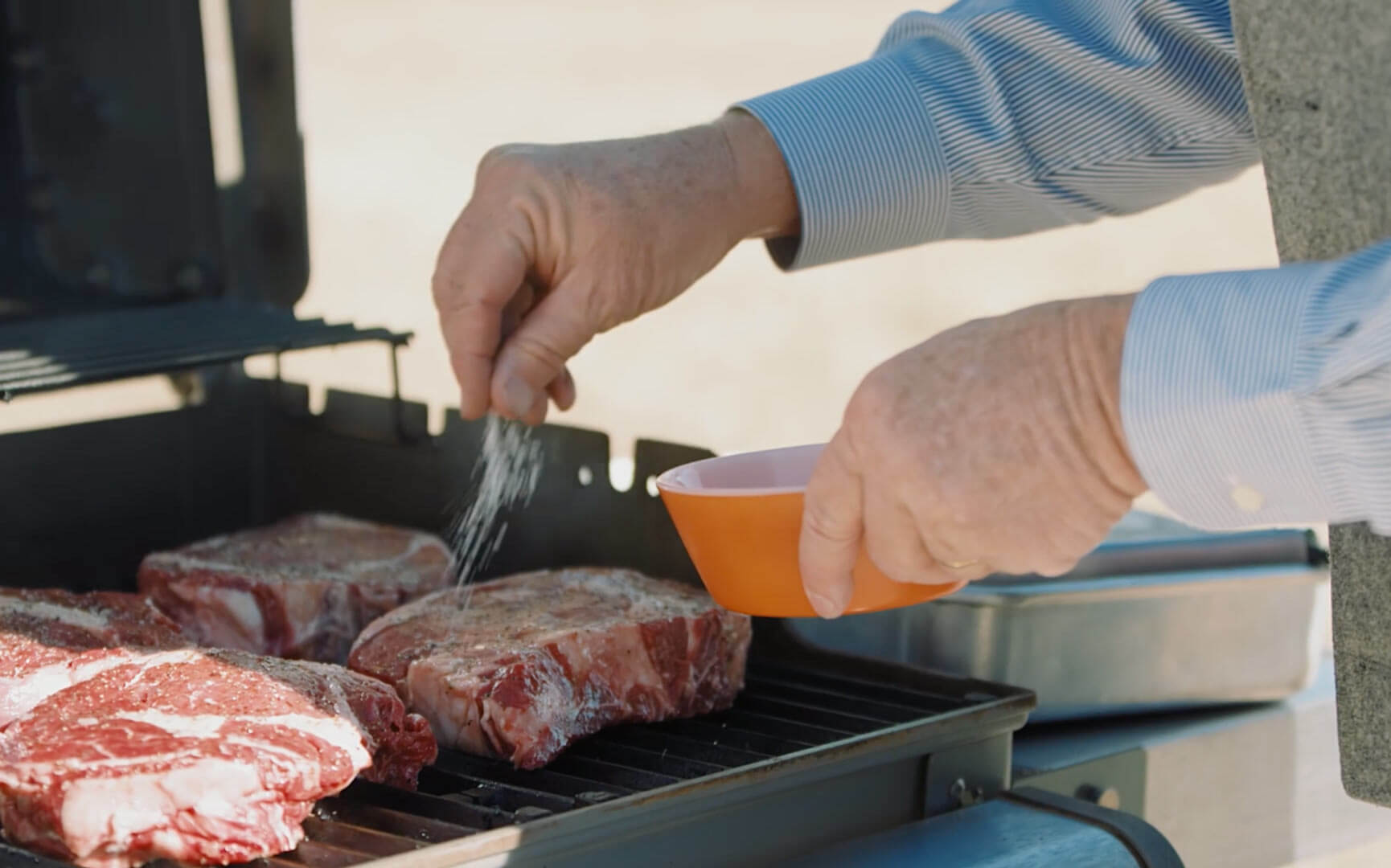 Hands sprinkling salt onto steaks on an outdoor grill.