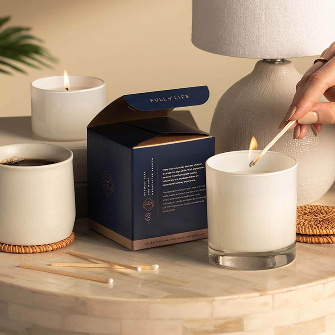 Hand lighting a Trapp Fragrances candle sitting on a tabletop with a lamp, matches, coffee mug and the candle package.