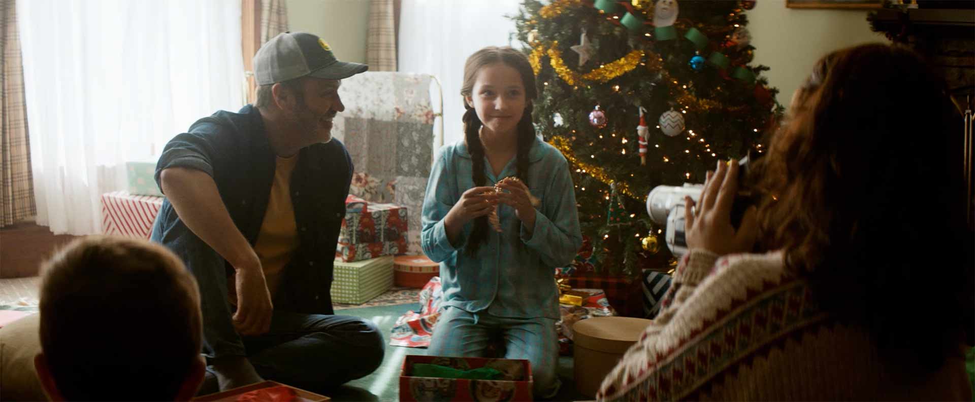 Young girl (about 10) kneels on the floor opening a present with her father watching her from the side.