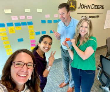 Selfie image of Amanda Hembree with John Deere employees at a training session in front of a wall of post-it notes