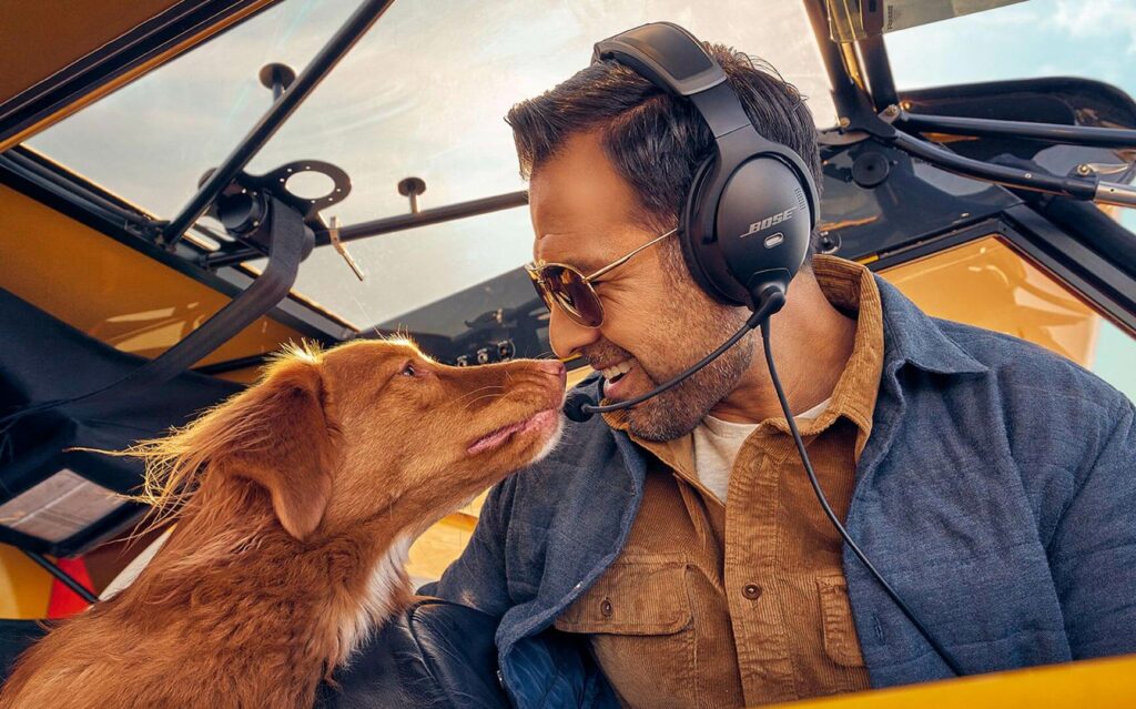 Male Pilot in small cockpit wearing Bose A30 headset smiling at his dog next to him