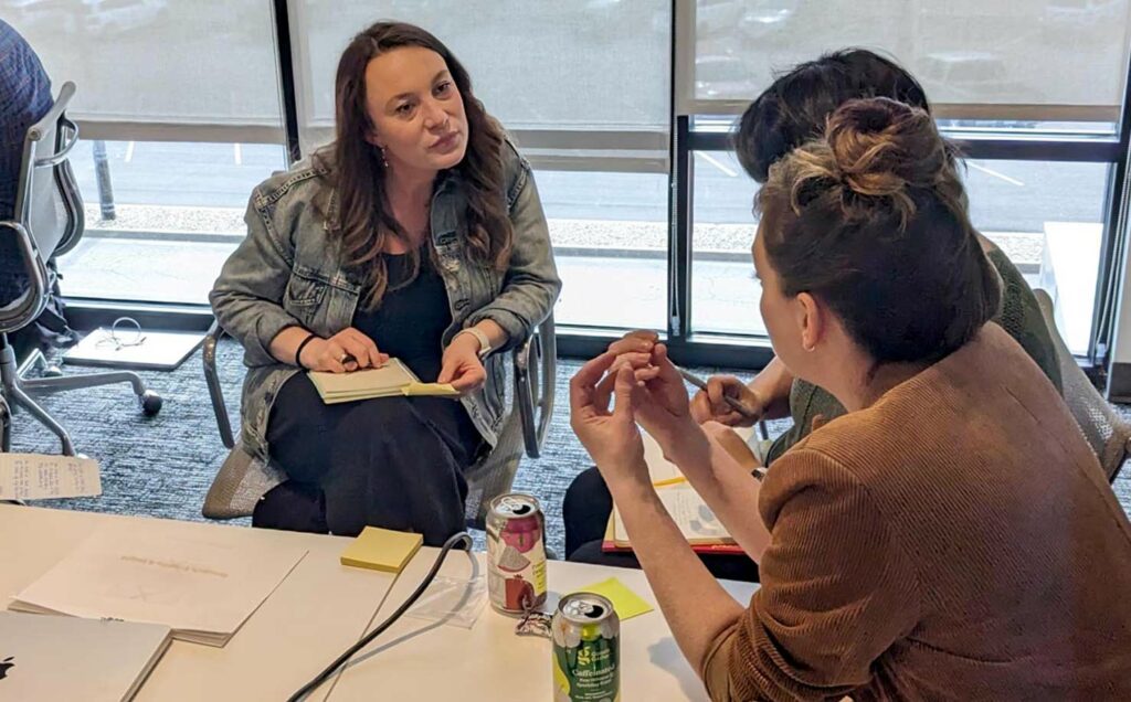 Several women sitting at corner of a conference room table leaning in and intently listening as the speaker gestures with her hands.